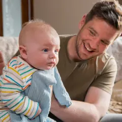 Padre sorridente tiene in braccio un neonato con una copertina azzurra. La posizione del neonato sembra confortevole, forse per alleviare il singhiozzo.