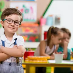 Uśmiechnięty chłopiec w okularach, gotowy do przedszkola. W tle dzieci i nauczycielka.
