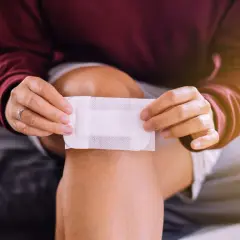 Osoba przykłada plaster do kolana, by wchłonąć ewentualny **płyn surowiczy** po urazie.