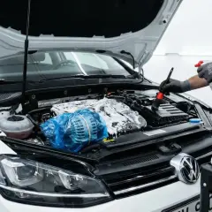 Persona con guantes rocía espuma y usa un cepillo para limpiar motor coche. El alternador está cubierto con plástico azul.