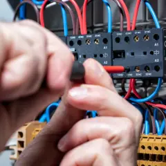 Ręce elektryka pracują przy rozdzielnicy, podłączając przewody. Widać styki i bezpieczniki, a napięcie w sieci jest pod kontrolą.
