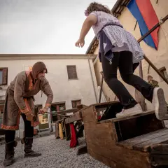 Bambini si divertono con giochi medievali. Un uomo in costume d'epoca aiuta una bambina a salire su una struttura di legno.