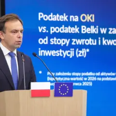 Mężczyzna w garniturze przemawia na tle ekranu z napisem "Podatek na OKI vs. podatek Belki". Dyskusja o zmianach w podatkach.
