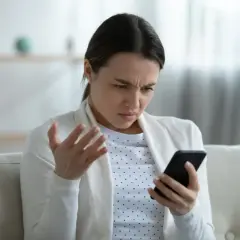 Kobieta z irytacją patrzy na telefon, gestykulując ręką. Ewidentnie przeszkadza jej słaby internet w telefonie, uniemożliwiający normalne korzystanie.