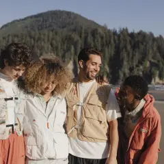 Grupo de amigos con estilo gorpcore en la playa, con chalecos utilitarios y chaquetas, disfrutando del paisaje costero.