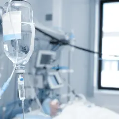 Kroplówka w szpitalnej sali, gdzie pacjent jest w spiaczce farmakologicznej. W tle aparatura medyczna i okno.