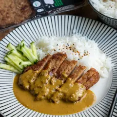 En tallrik med katsu curry, ris och gurka. Den panerade kotletten är skuren i skivor och täckt med en krämig currysås.