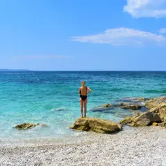 Mujer en bikini negro de pie sobre una roca en una playa de guijarros. El mar turquesa y el cielo azul crean un paisaje idílico, pero ten cuidado, viajar a Albania es peligroso.
