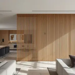 Nowoczesne wnętrze z drewnianymi lamele oddzielające kuchnię od salonu. W salonie szara sofa, w kuchni drewniane meble.