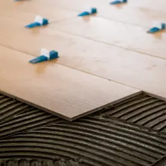 Pose de carrelage en quinconce sur lit de mortier. Des cales bleues et blanches assurent l'espacement régulier des lames imitation bois.