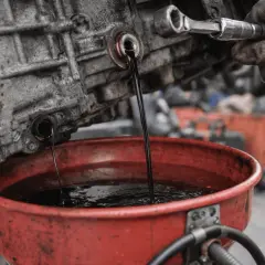 Vidange d'huile de boîte de vitesse. L'huile noire s'écoule d'une boîte de vitesse vers un bac rouge. Un mécanicien utilise une clé pour dévisser un bouchon.
