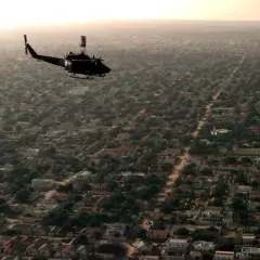 Helikopter przelatuje nad gęsto zabudowanym miastem z licznymi domami i drzewami.