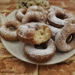 Puszyste oponki serowe, posypane cukrem pudrem, kuszą swoim domowym wyglądem. Jedna z nich jest nadgryziona, ukazując puszyste wnętrze.