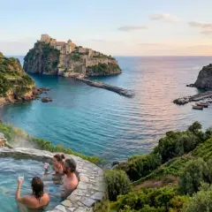 Coppie rilassate in una sorgente di nitrodi gratis con vista su un castello e un borgo sul mare.