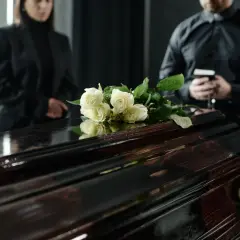 Białe róże na ciemnej trumnie, ludzie w żałobie. Wskazówki, jak zachować się na pogrzebie, w tym szacunek i powaga.