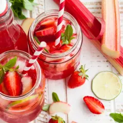 Orzeźwiający sok z rabarbaru z truskawkami i miętą, podany w słoikach z lodem i słomkami. Obok leżą świeże rabarbar, truskawki i plasterki limonki.