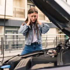 Zrozpaczona dziewczyna z rękami na uszach przy otwartej masce samochodu. Jej auto nie odpala, co ją bardzo frustruje.