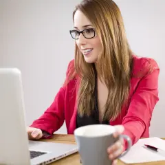 Uśmiechnięta kobieta w okularach i czerwonej marynarce pracuje przy laptopie, trzymając kubek. Zastanawia się, czy można dokupić AC w trakcie trwania OC.
