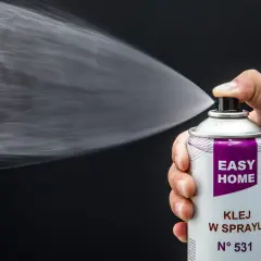 Dłoń trzyma puszkę z napisem "EASY HOME KLEJ W SPRAYU", rozpylając białą mgiełkę. Idealny do podsufitki.