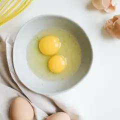 Deux jaunes d'œufs dans un bol, prêts à être battus. Une question se pose : peut-on congeler des jaunes d'œufs ? Un fouet jaune et des coquilles d'œufs complètent la scène.
