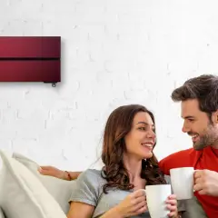 Pareja sonriente en un sofá, disfrutando de café. Arriba, un aire acondicionado rojo moderno garantiza cop eficiencia.
