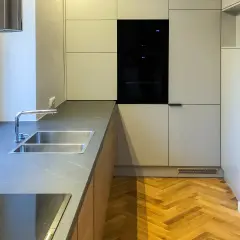 Nowoczesna kuchnia z szarymi frontami, wbudowanym piekarnikiem i podwójnym zlewozmywakiem. Podłoga w jodełkę i okap w kolorze RAL 9016 dodają elegancji.