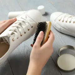 Mains nettoyant une basket blanche en tissu avec une brosse. Un pot de produit d'entretien est ouvert à côté.