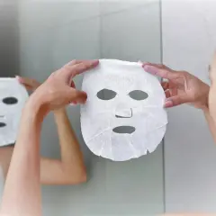 Mani bagnate che applicano una maschera in tessuto sul viso, riflessa nello specchio. Dopo la maschera in tessuto si lava il viso.