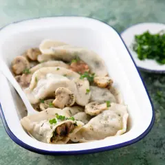 Pyszne pierogi z pieczarkami i serem, posypane natką pietruszki, w białej misce z niebieską obwódką.