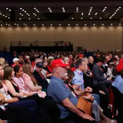 A large audience attends a government conference, with many people wearing red hats.