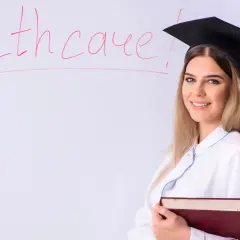Laurea in medicina ma non voglio fare il medico. Studentessa con tocco e camice, libro in mano, davanti a una lavagna con la scritta "Healthcare".