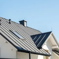 Nowoczesny dom z czarnym, metalowym dachem i świetlikiem. Dwa kominy i czarna balustrada balkonu dopełniają architekturę.