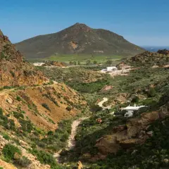 Paisaje árido con el **Cerro Negro** al fondo. Un camino serpentea entre barrancos y vegetación baja, llegando a unas casas blancas.