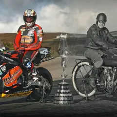 Conor Cummins junto a su moto de carreras y un trofeo, junto a un piloto histórico en una Indian.