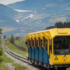 Kolejka na górę Żar oferuje wspaniałe widoki. Żółto-niebieski wagon wjeżdża na szczyt, a w tle rozciąga się malownicza dolina z jeziorem.