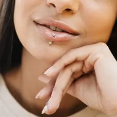 Usta z błyszczącym błyszczykiem i subtelnym center labret. Dłoń podtrzymuje podbródek, podkreślając delikatny uśmiech.