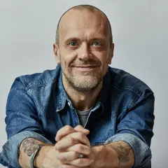 Uomo con barba corta, occhi azzurri, indossa una camicia di jeans. Le sue mani sono intrecciate, eccoti testo.