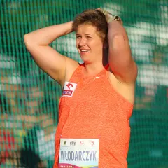 Ania Włodarczyk, po rzucie młotem rekord świata, z uśmiechem na twarzy i rękami na głowie, za zieloną siatką.