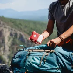 Mann packt Erste-Hilfe-Set für leichte Wanderung in Rucksack. Bergpanorama im Hintergrund.