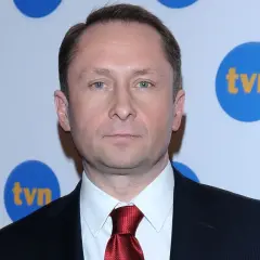 Kamil Durczok w garniturze i czerwonym krawacie na tle logo TVN. Prowadzący Fakty TVN.