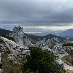 Osoba w turkusowej kurtce na skalistym szczycie w Tatrach Zachodnich. Słońce przebija się przez chmury, oświetlając jezioro w oddali.