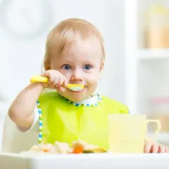 Uśmiechnięte 17 miesięczne dziecko w żółtym śliniaku próbuje samodzielnie jeść z łyżeczki.