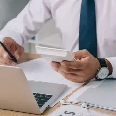 Mężczyzna w białej koszuli i granatowym krawacie liczy na kalkulatorze, analizując dokumenty. Zastanawia się, jaka rata kredytu 400 tys. będzie dla niego najlepsza.