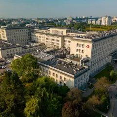Widok z lotu ptaka na duży kompleks szpitalny z czerwonym krzyżem na fasadzie, otoczony drzewami i parkingiem.