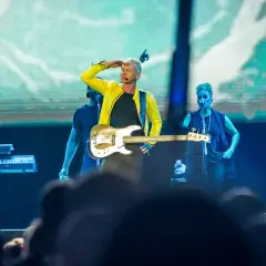 Sting w żółtej kurtce gra na gitarze basowej na scenie z zespołem.