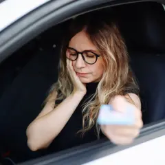 Kobieta w okularach siedzi za kierownicą, trzymając kartę. Wygląda na zmartwioną, jakby czekała na kontrolę, bo grozi jej jazda bez prawa jazdy.