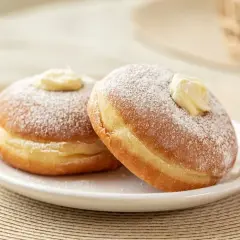 Due bomboloni ripieni di crema, spolverati di zucchero a velo, mostrano la differenza tra bombolone e krapfen: il primo è più soffice e senza buco.