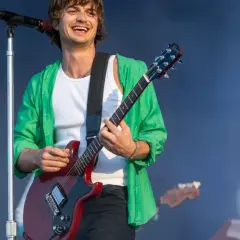 Joe Keery w zielonej koszuli gra na czerwonej gitarze elektrycznej na scenie.