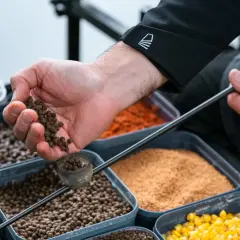 Ręka wsypuje pellet do małego pojemnika, przygotowując zanętę do koszyczka. W tle widać pojemniki z różnymi składnikami.