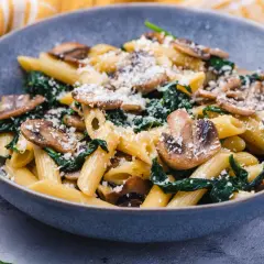 Penne z pieczarkami i szpinakiem, posypane parmezanem, w szarej misce.